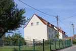 Ein Haus mit einem Zaun und einer Straße, umgeben von seitlich sichtbaren Wohnblöcken.